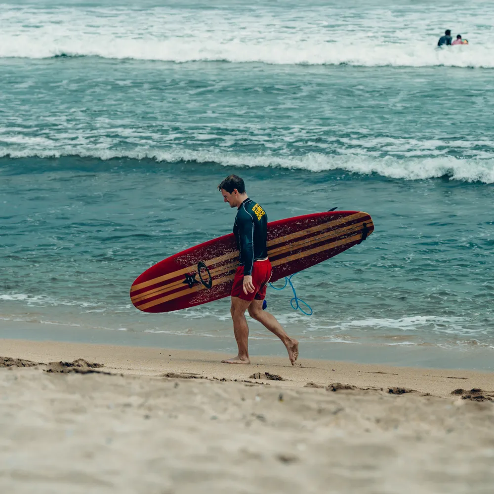 A surfer carrying a red board along the shore